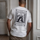 A man viewed from behind wearing a white t-shirt with a large black graphic print on the back. The graphic features the text 'PARADAIZA' and 'THE CARD OF CREATION, ABUNDANCE, AND GROWTH' with an illustration.