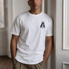 A man wearing a white crewneck t-shirt with a black graphic on the left chest, leaning against a white paneled door in an indoor setting.