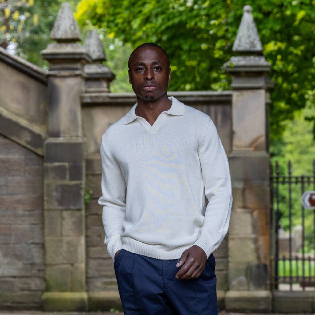 Man wearing a ivory paradaiza sweater and dark pants standing in front of a stone wall with greenery.