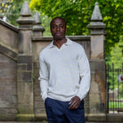 Man wearing a ivory paradaiza sweater and dark pants standing in front of a stone wall with greenery.