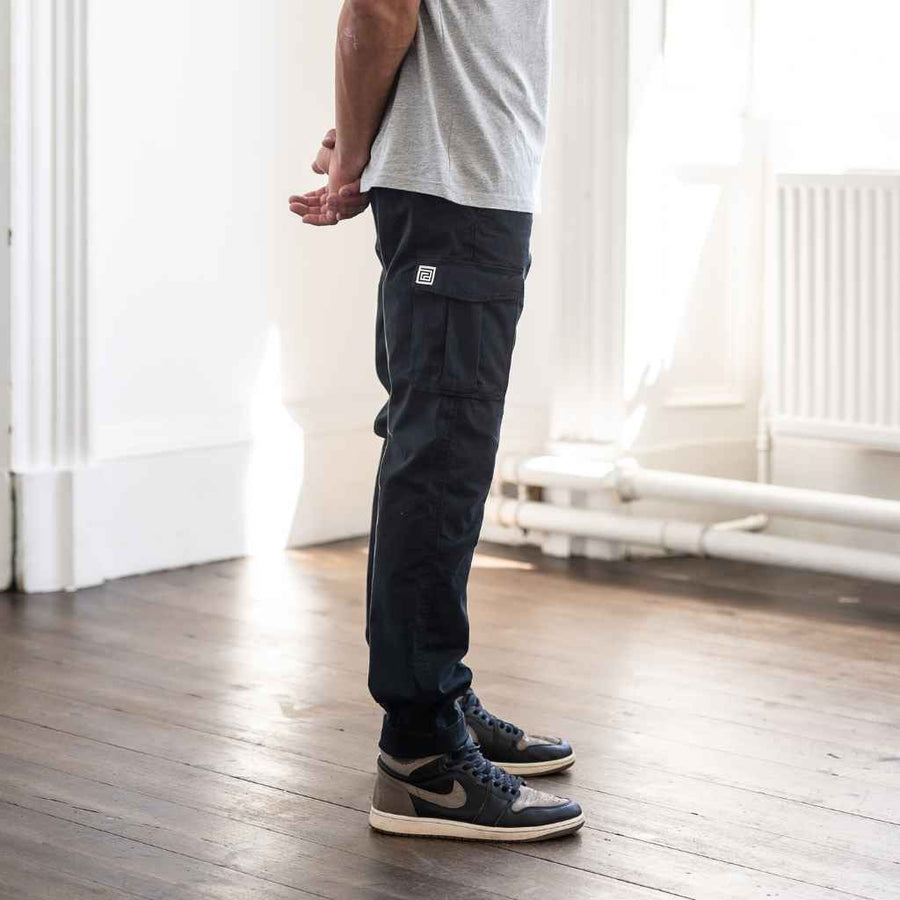 A person standing in an indoor setting wearing paradaiza black cargo pants and a grey shirt with sneakers.