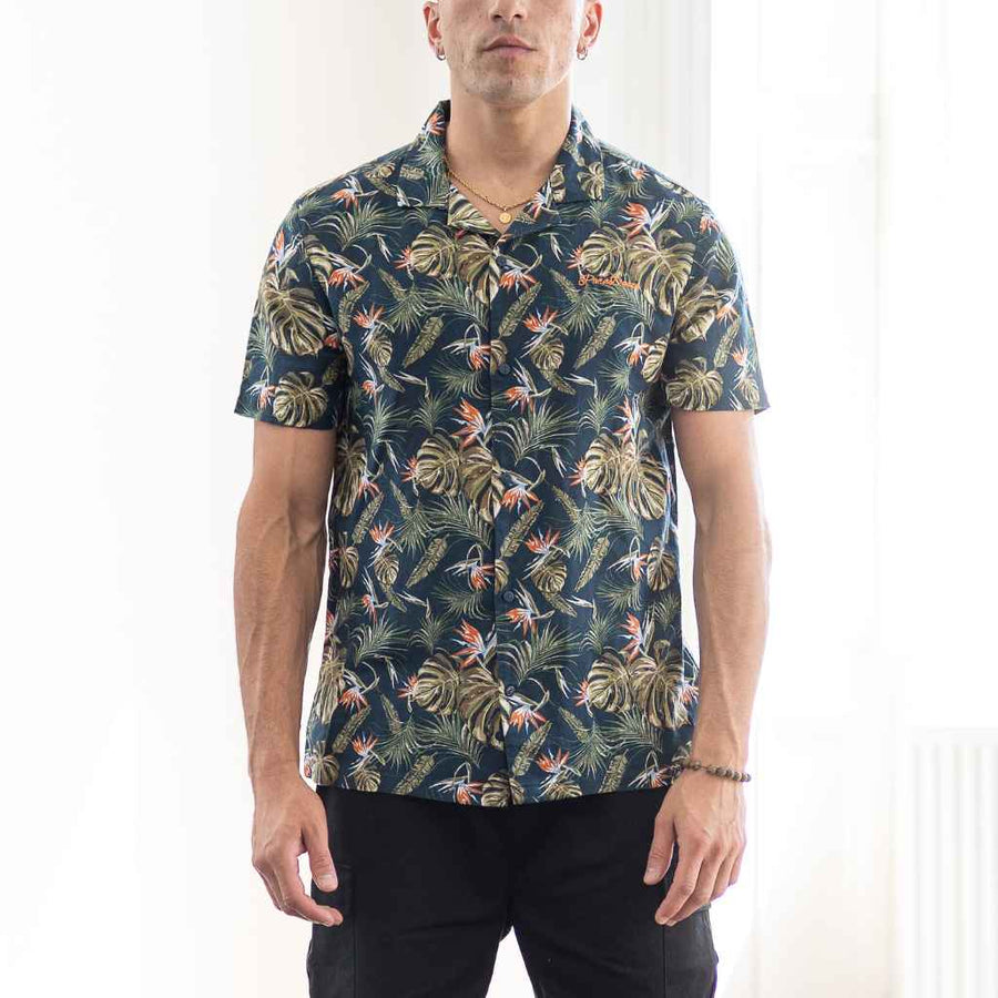 A male model wearing a Para daiza navy shirt with a tropical leaf and fruit print, paired with dark trousers.