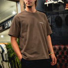 A man wearing a brown crewneck t-shirt with a small white embroidered logo on the left chest, standing in a retail store environment.