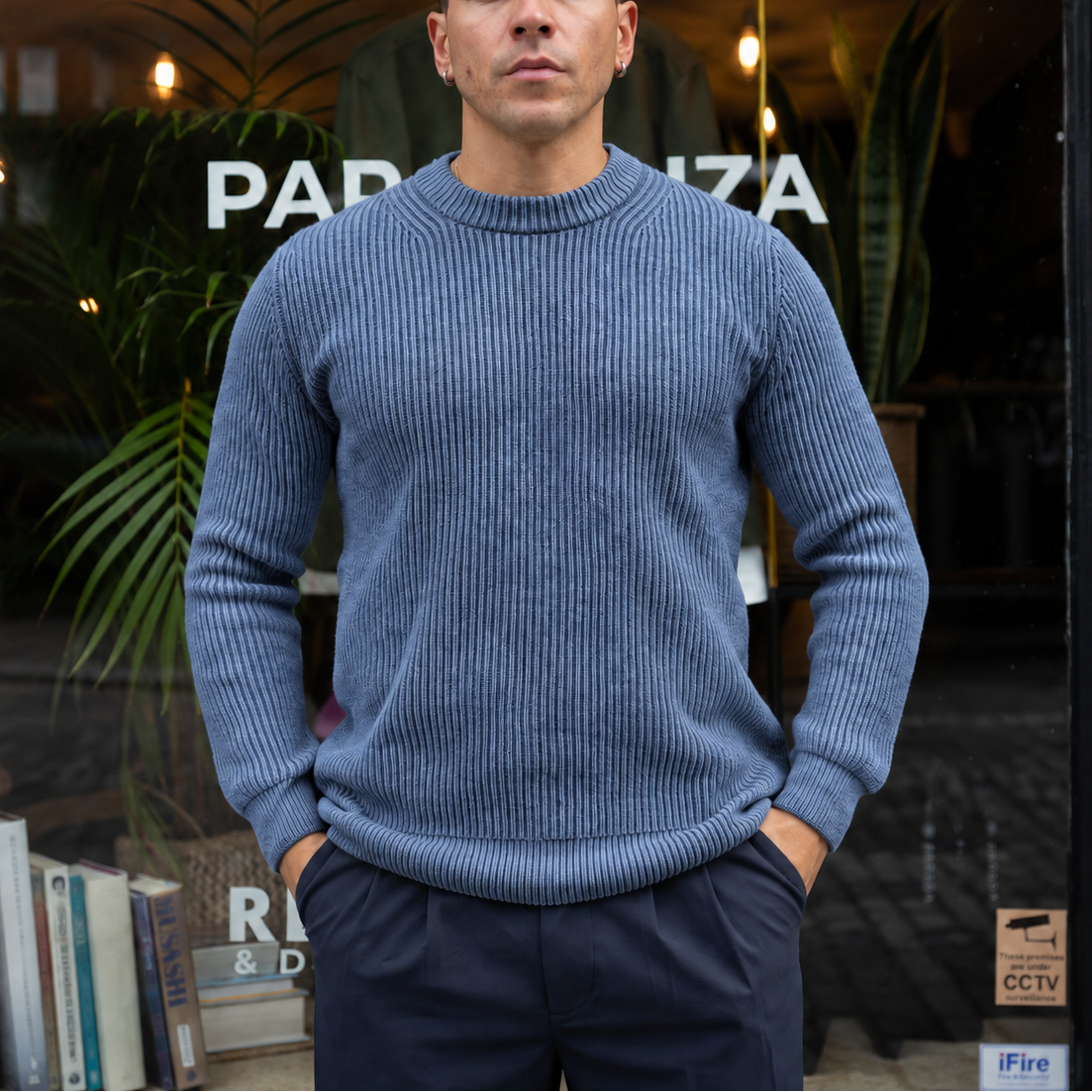A man wearing a blue ribbed knit crewneck sweater and dark trousers stands in front of a storefront window with plants and books visible.