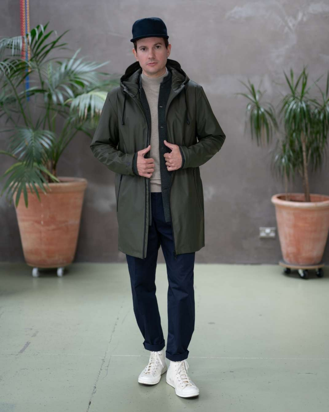 Person wearing a paradaiza green coat, dark cap, and white sneakers standing indoors with plants in the background