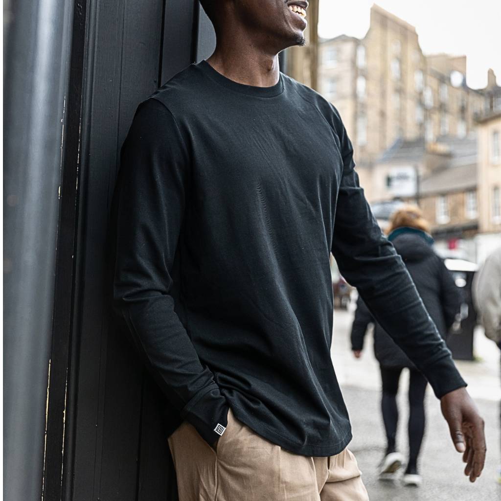 Man wearing a paradaiza black long-sleeve shirt leaning against a wall on a city street.
