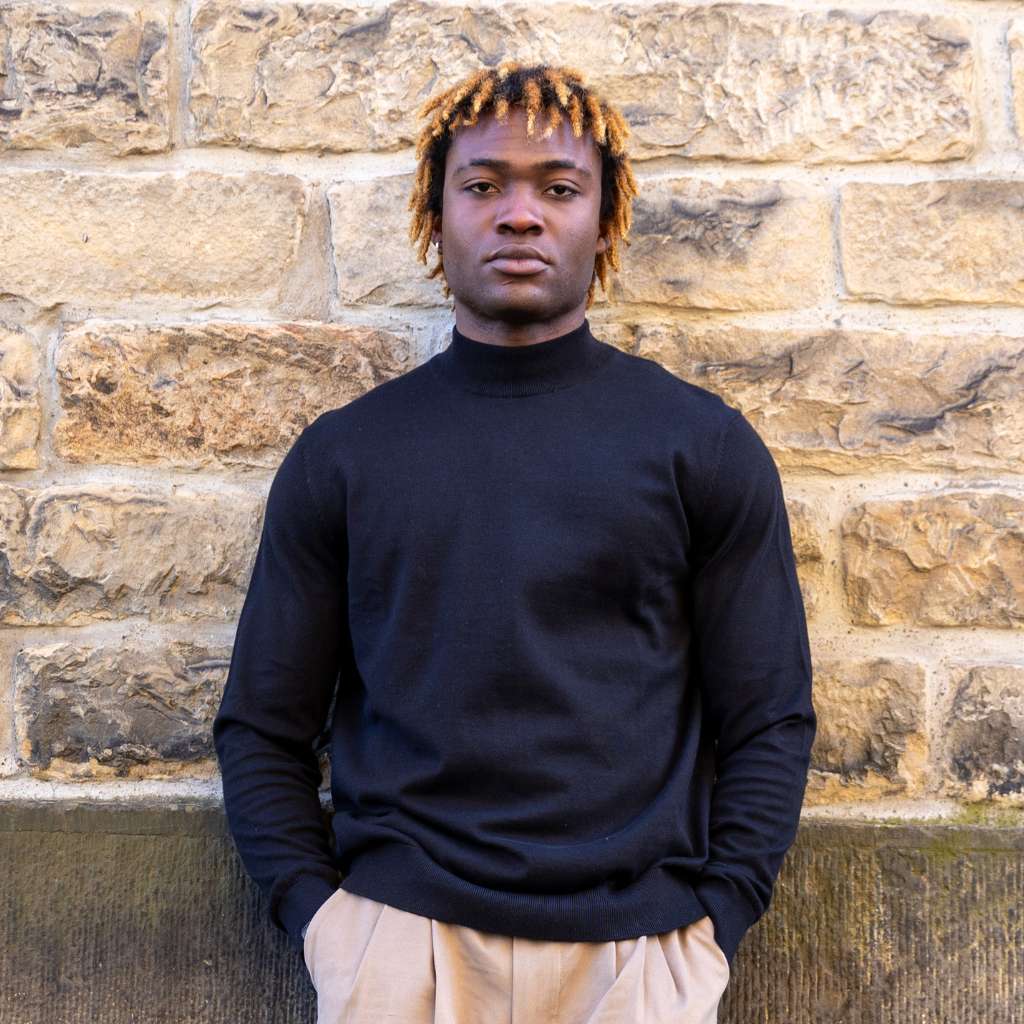 Person wearing a paradaiza black turtleneck sweater and beige pants against a stone wall.