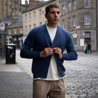 A man wearing a navy blue cardigan over a white t-shirt and beige trousers, standing on a cobblestone street in an urban setting.