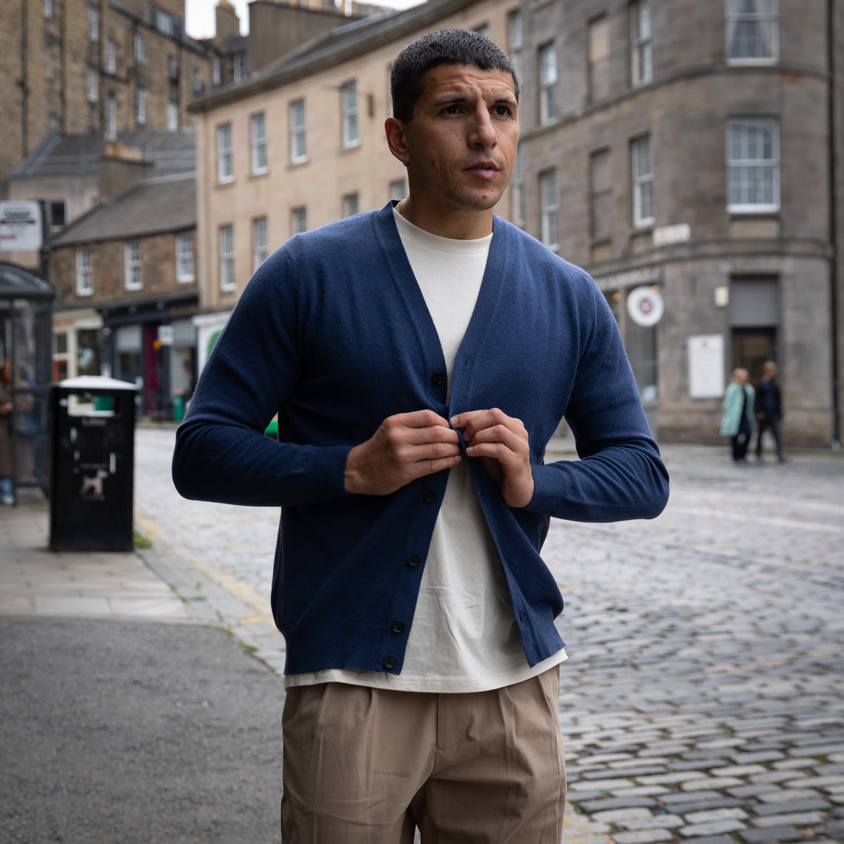 A man wearing a navy blue cardigan over a white t-shirt and beige trousers, standing on a cobblestone street in an urban setting.
