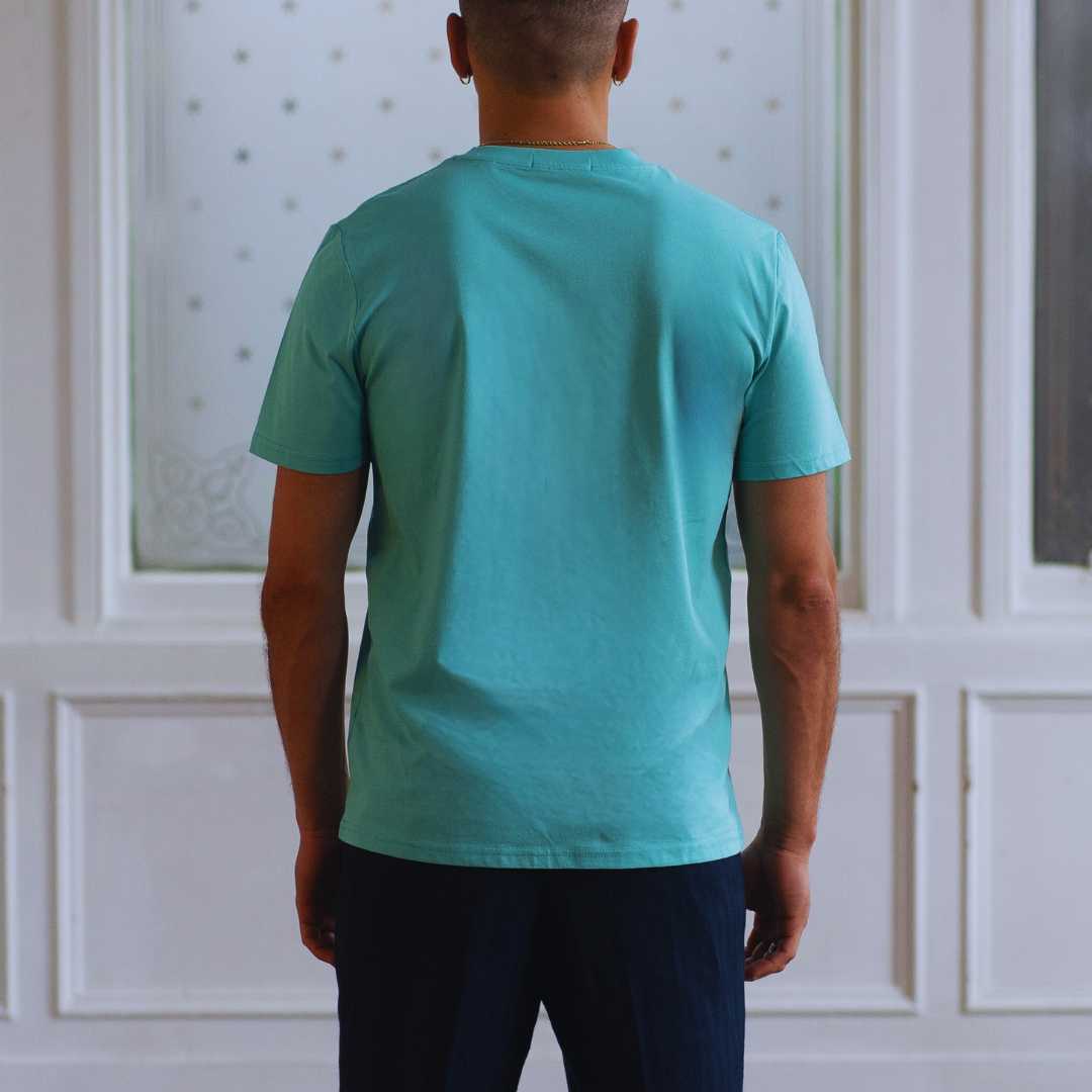 Man wearing a  paradaiza teal t-shirt with a small logo against a white paneled wall.
