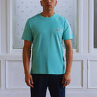 Man wearing a  paradaiza teal t-shirt with a small logo against a white paneled wall.