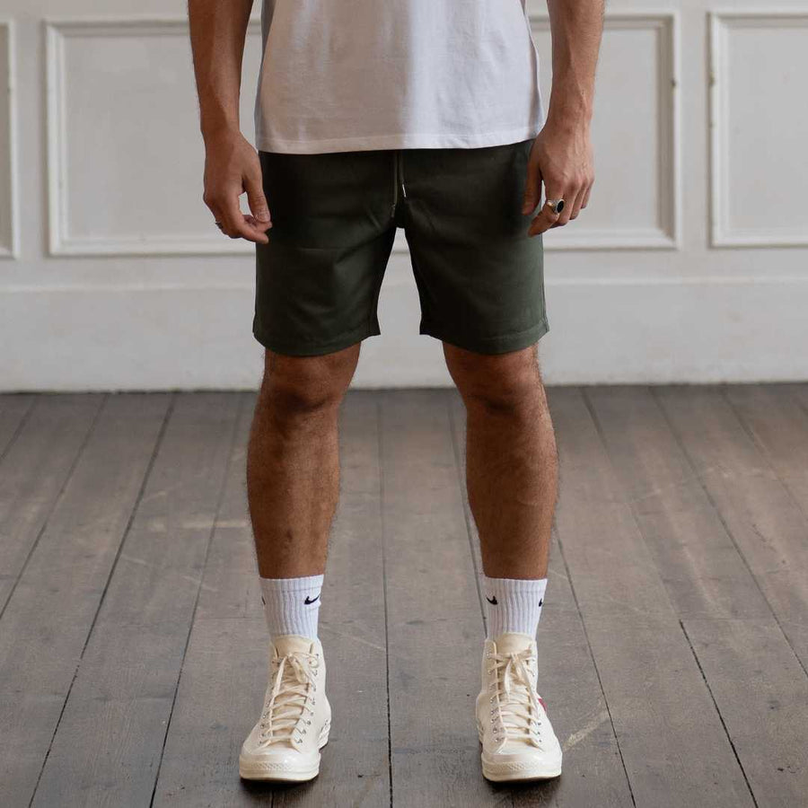 Person wearing green paradaiza shorts, white socks, and beige sneakers in a room with wooden flooring and white paneled walls.