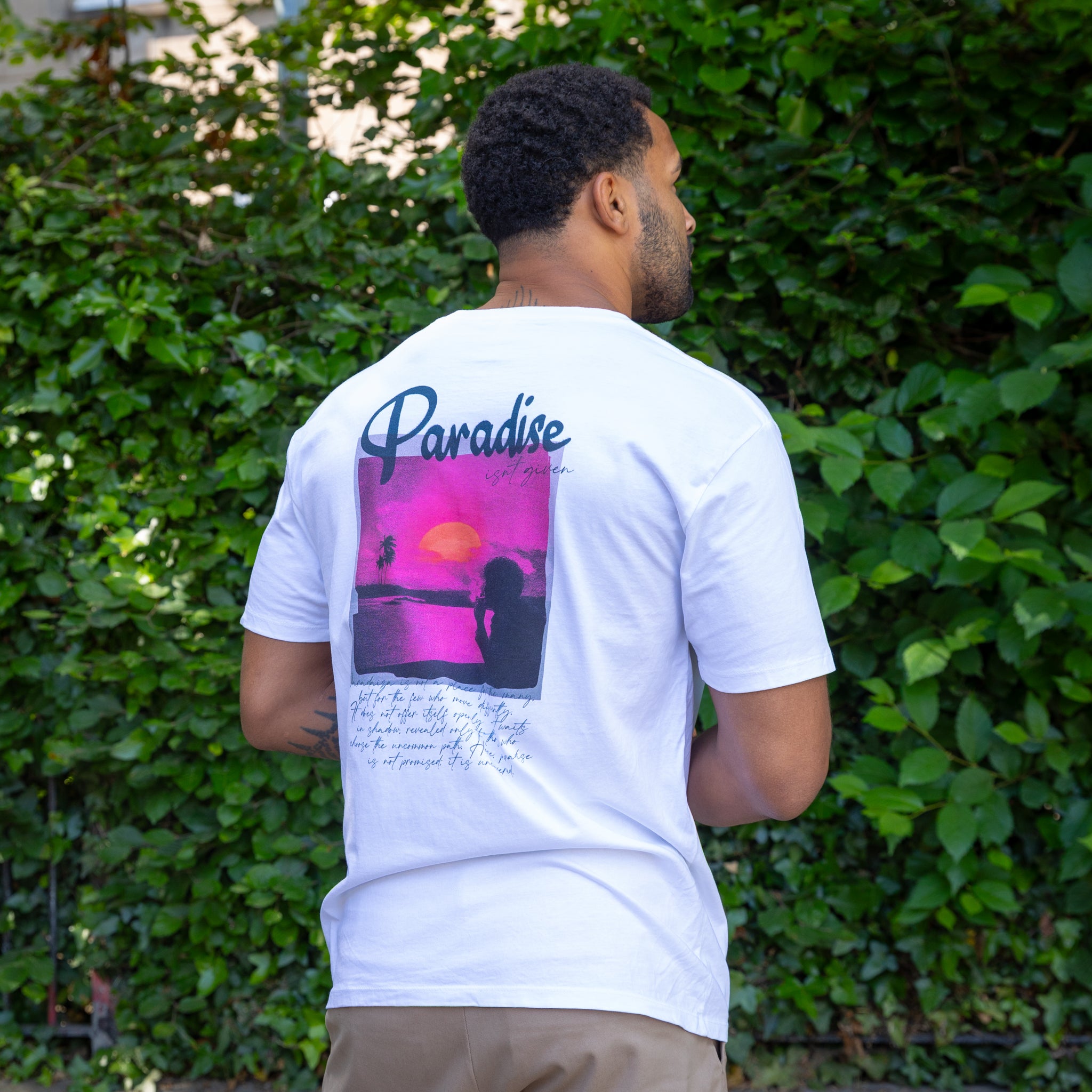 Man wearing a paradaiza white t-shirt with a colorful graphic on the back, standing in front of green foliage.
