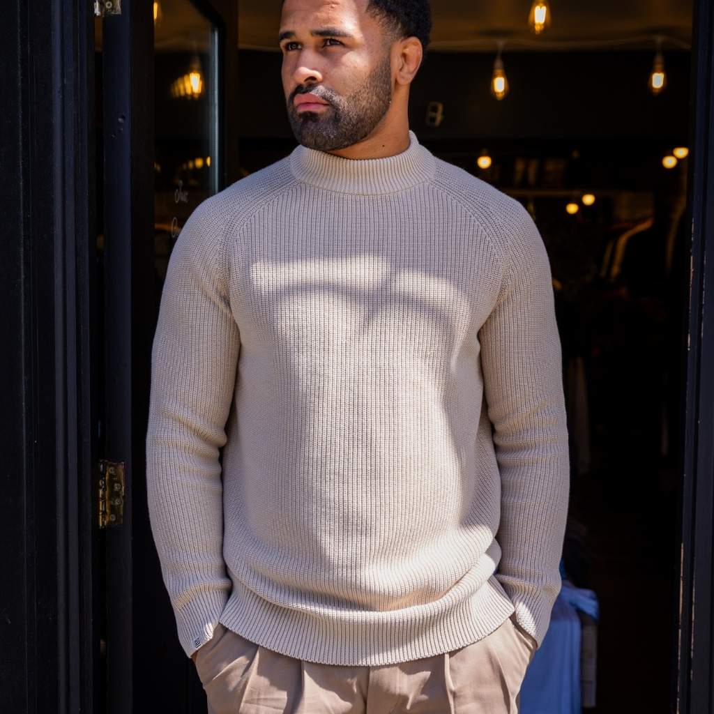 Man wearing a  paradaiza beige sweater standing in front of a dark doorway with lights.