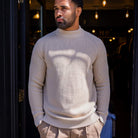 Man wearing a  paradaiza beige sweater standing in front of a dark doorway with lights.