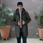 A male model wearing a long, Paradaiza khaki-colored parka jacket with a hood, paired with a white t-shirt and beige pants.