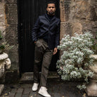 Model  wearing a dark denim paradaiza  jacket and pants with a cap, standing indoors with plants in the background.