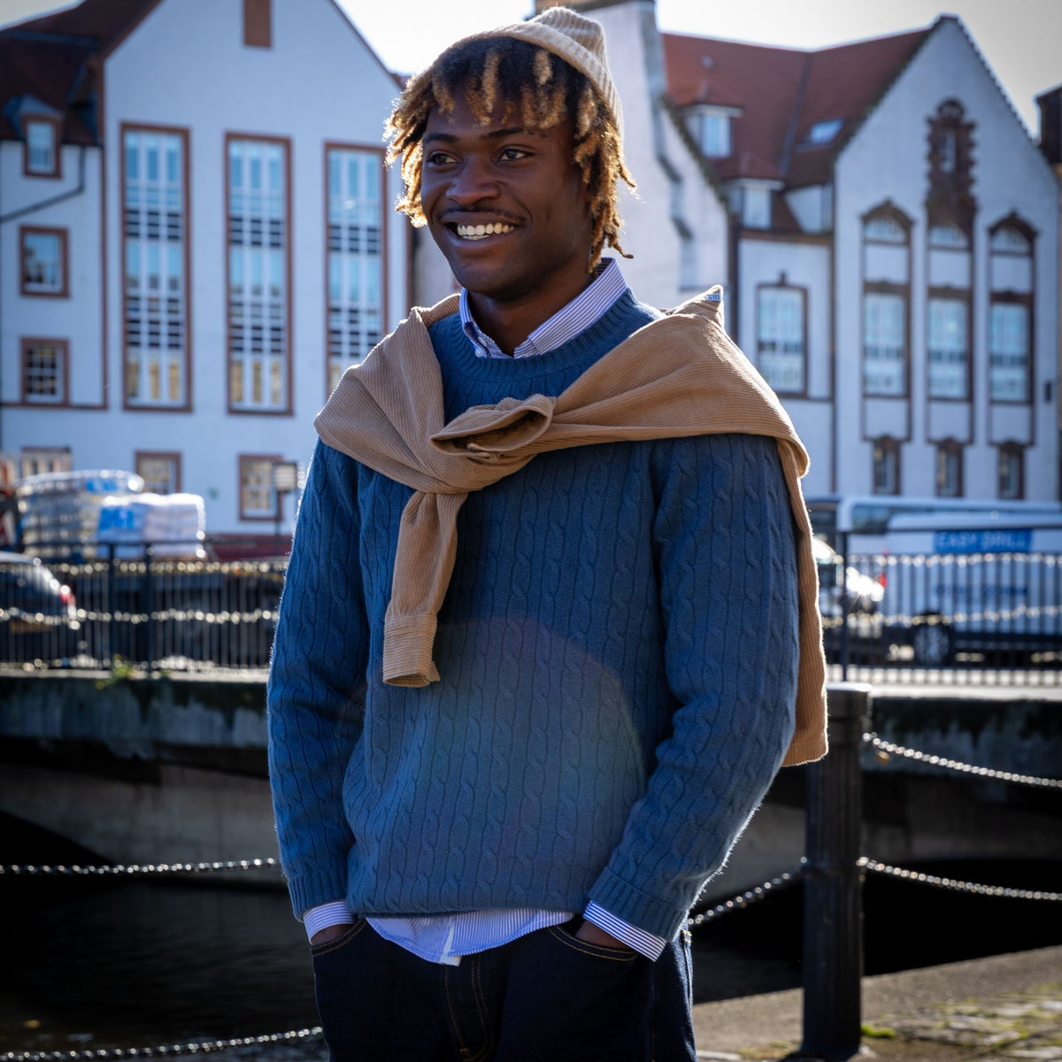 A smiling man standing outdoors in front of white buildings, wearing a blue cable-knit sweater over a white collared shirt, with a tan scarf draped around his shoulders and a beige beanie on his head.