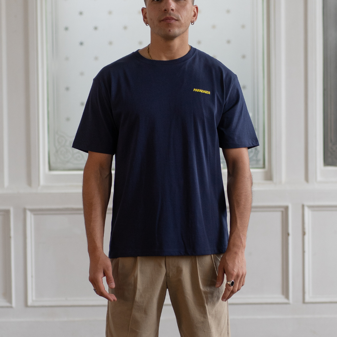 model  wearing a  paradaiza navy blue t-shirt with a  yellow logo in front of a white paneled wall.