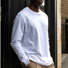 Person wearing a paradaiza white long-sleeve shirt and beige pants standing against a dark door.