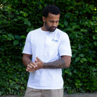 Man wearing a paradaiza white t-shirt with a logo standing against a green leafy background
