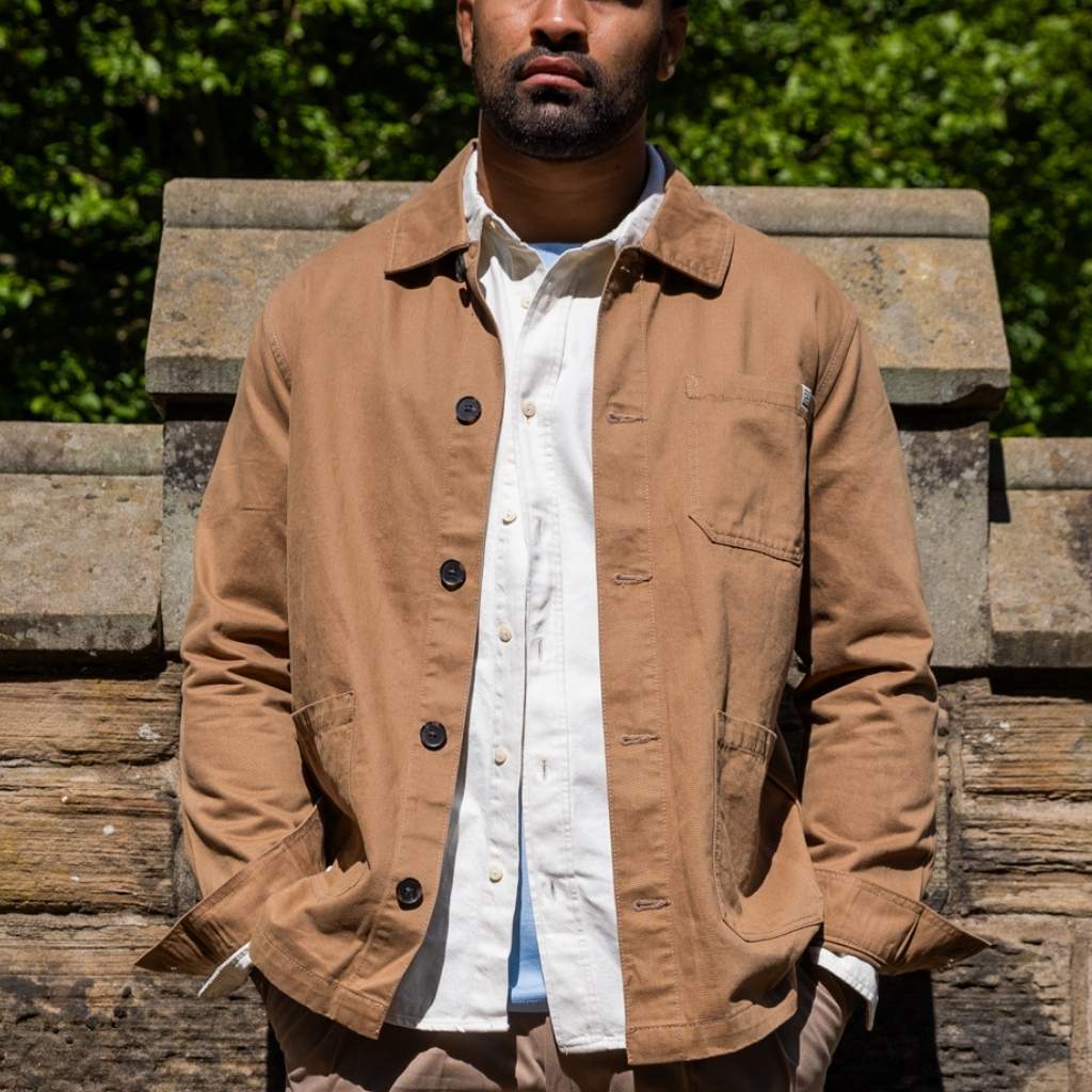 Man wearing a tan paradaiza worker  jacket over a white shirt with a blurred green background