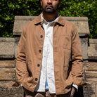 Man wearing a tan paradaiza worker  jacket over a white shirt with a blurred green background