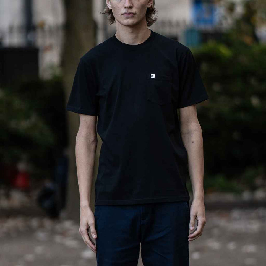 A person wearing a paradaiza black crew neck t-shirt with a small logo on the chest and a small brand flag on the sleeve.