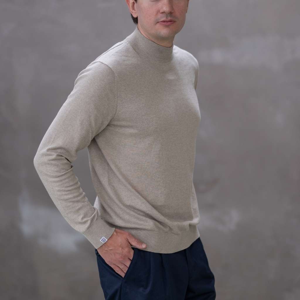 Man wearing a paradaiza beige turtleneck sweater against a gray 