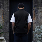 Man wearing a paradaiza black vest over a white shirt standing in front of a stone wall.