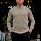 A man wearing a beige ribbed knit crewneck sweater and dark pants, standing in front of a dark storefront with plants and books visible in the background.