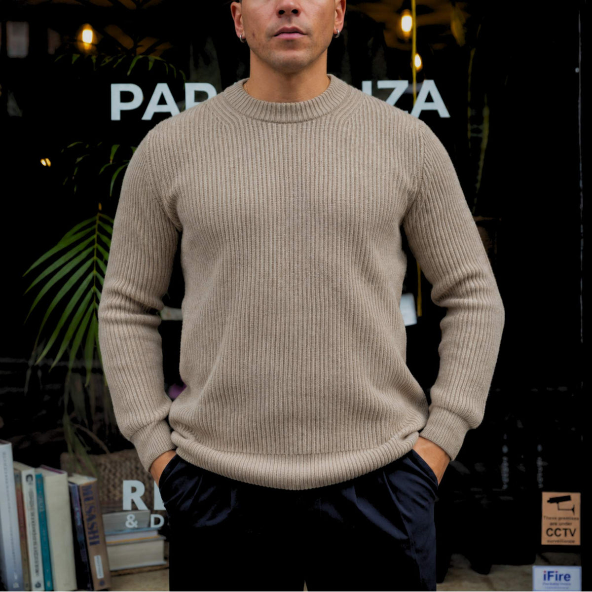 A man wearing a beige ribbed knit crewneck sweater and dark pants, standing in front of a dark storefront with plants and books visible in the background.