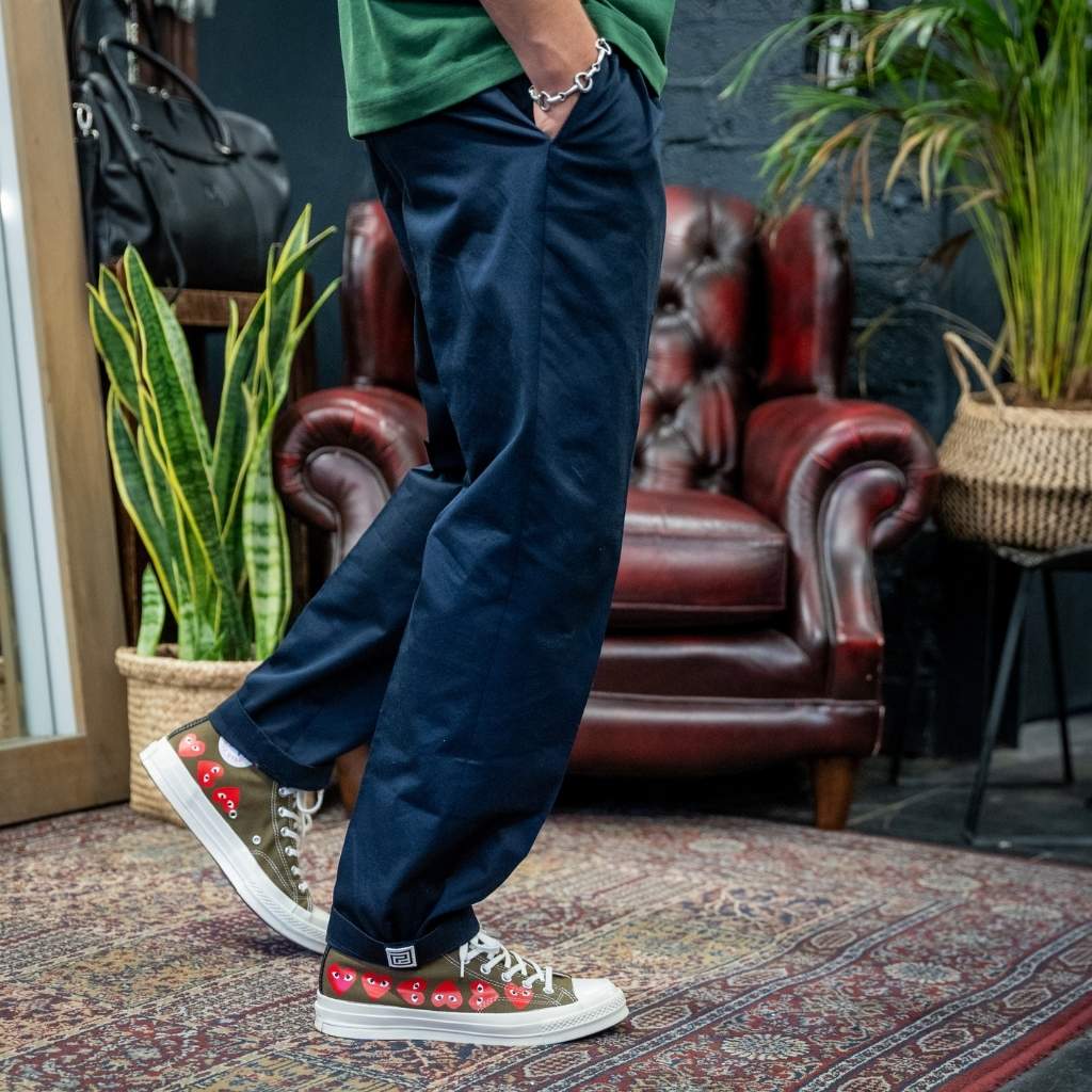 A person wearing dark navy blue relaxed-fit pants, a green t-shirt, and green low-top sneakers with red cartoon characters on the sides, standing on a patterned rug in front of a brown leather armchair and potted plants.
