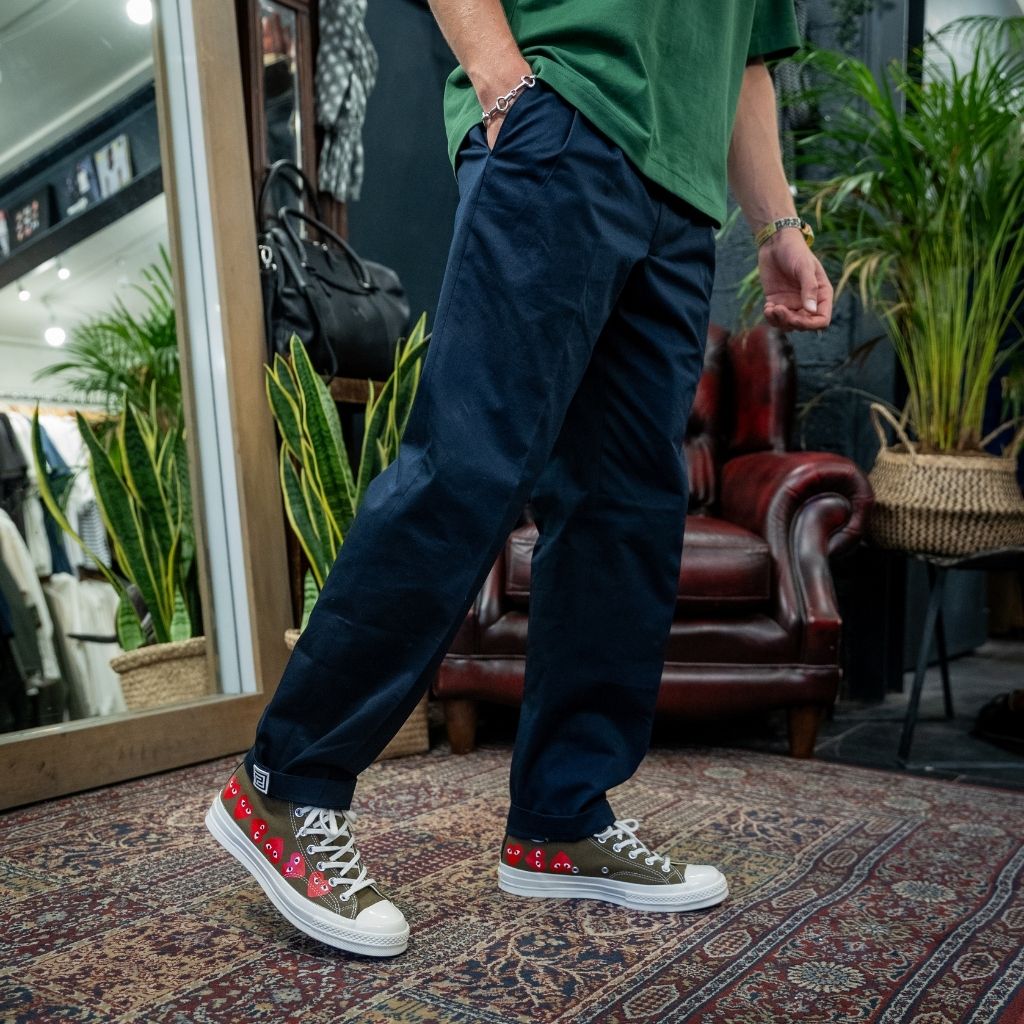 A person wearing dark navy blue relaxed-fit pants, a green t-shirt, and Converse sneakers with red floral embroidery, standing on a patterned rug in a store setting.