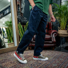 A person wearing dark navy blue relaxed-fit pants, a green t-shirt, and Converse sneakers with red floral embroidery, standing on a patterned rug in a store setting.