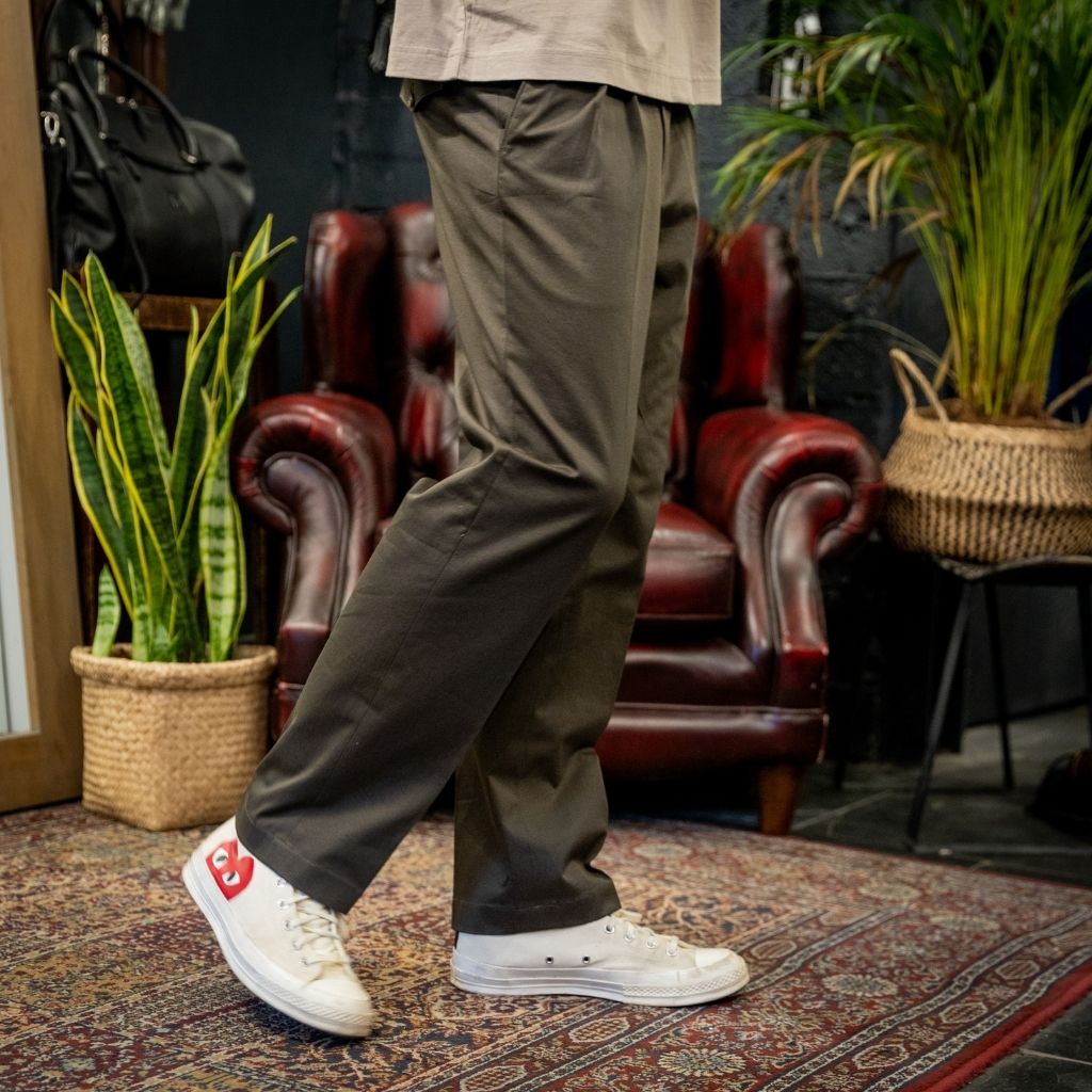 A person wearing loose olive green pants and white low-top sneakers with a red heel tab, standing on a patterned rug in a styled indoor room with a red leather armchair and potted plants.