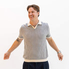 A smiling man wearing a paradaiza gray and white vertically striped short-sleeve knit polo shirt with cream-colored collar, sleeve cuffs, and hem, paired with dark navy trousers, set against a plain white studio background.