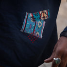 A close-up of a dark navy blue shirt featuring a colorful geometric patchwork design on the chest, with a hand wearing a silver ring visible in the lower right corner.