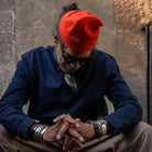 A man wearing a bright orange-red knit beanie, a dark blue button-up shirt, sunglasses, and multiple silver bracelets, sitting against a textured stone wall.