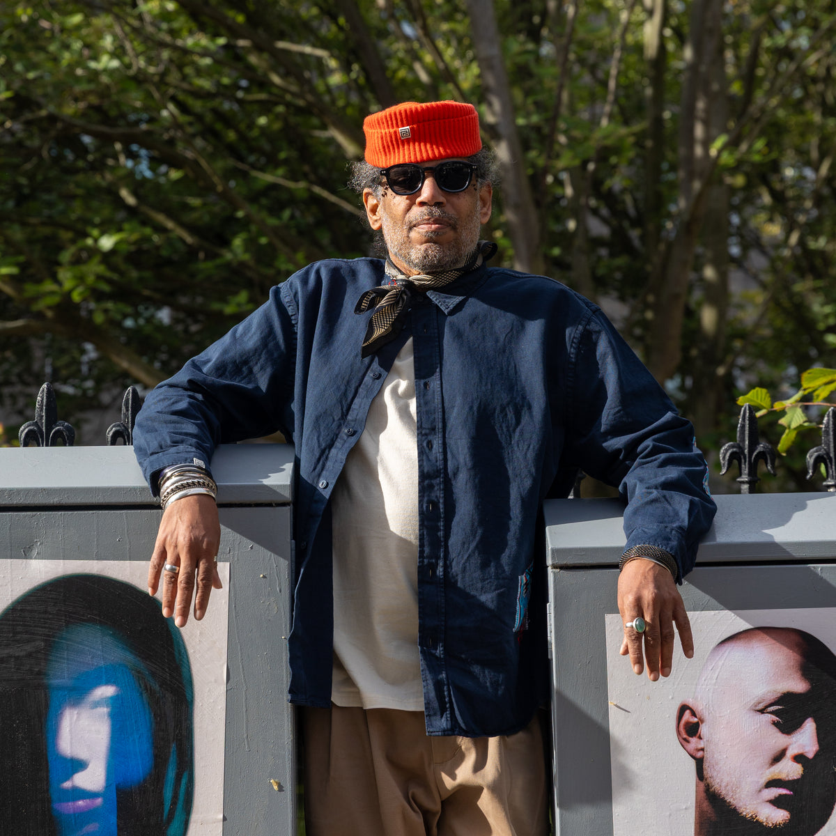 A man wearing a bright orange knit cap, black sunglasses, a navy blue shirt over a cream undershirt, and beige trousers leans against a gray metal barrier outdoors with trees in the background.