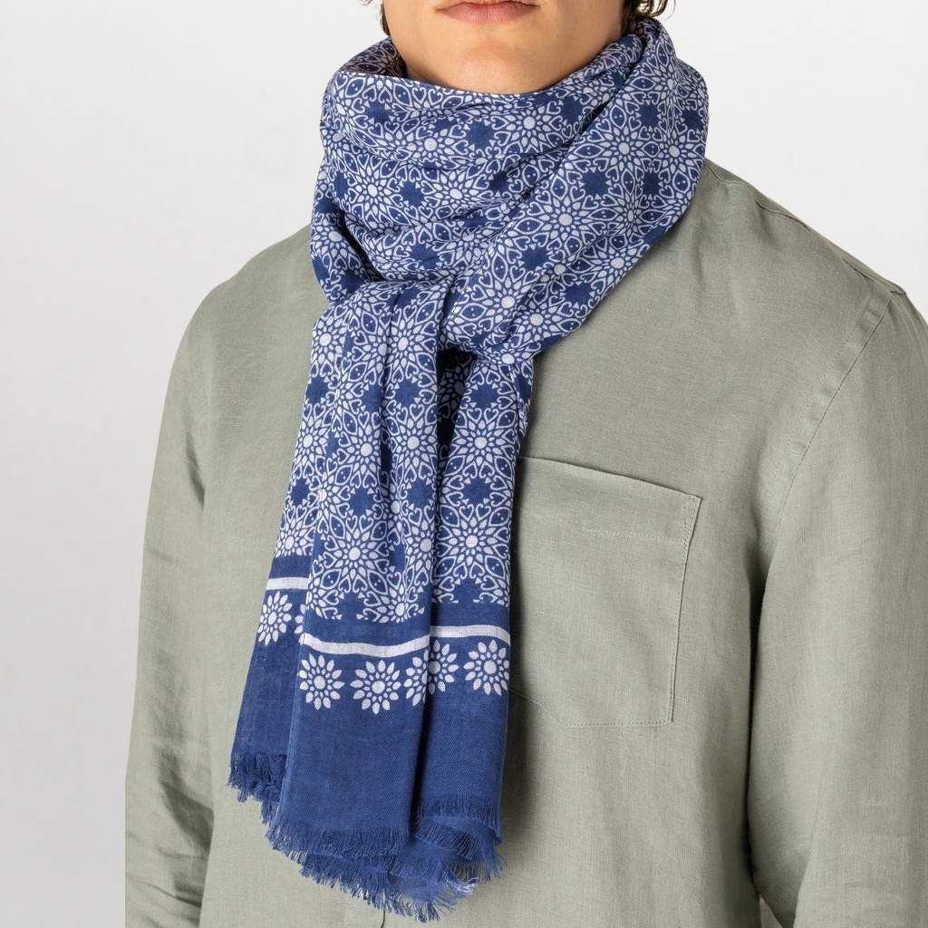 A person wearing a blue and white patterned scarf wrapped around the neck, paired with a light sage green shirt against a plain light gray background.