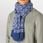 A person wearing a blue and white patterned scarf wrapped around the neck, paired with a light sage green shirt against a plain light gray background.