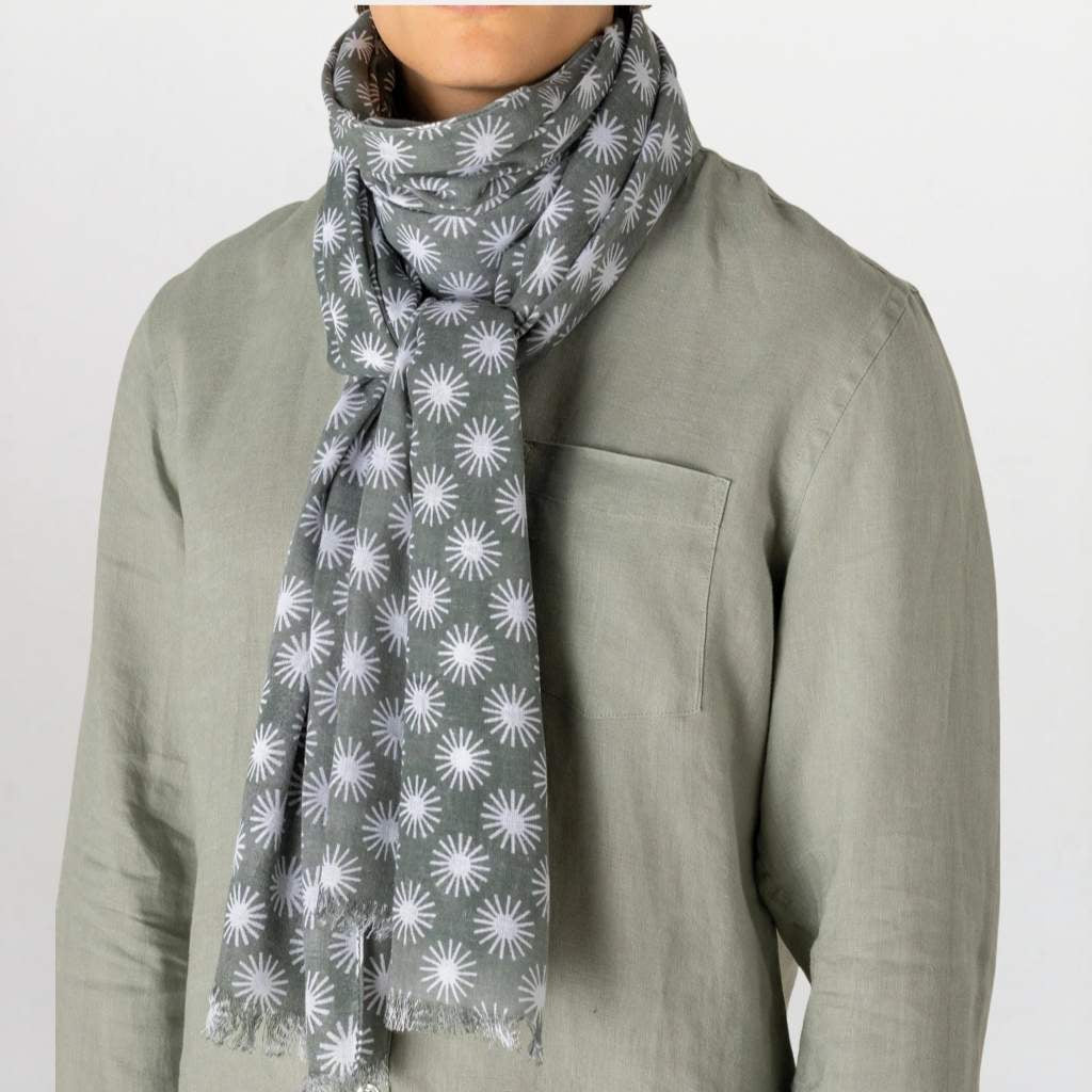 A gray scarf with a white repeating sunburst pattern, styled around the neck of a person wearing a sage green linen shirt against a plain light background.