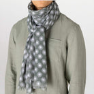 A gray scarf with a white repeating sunburst pattern, styled around the neck of a person wearing a sage green linen shirt against a plain light background.