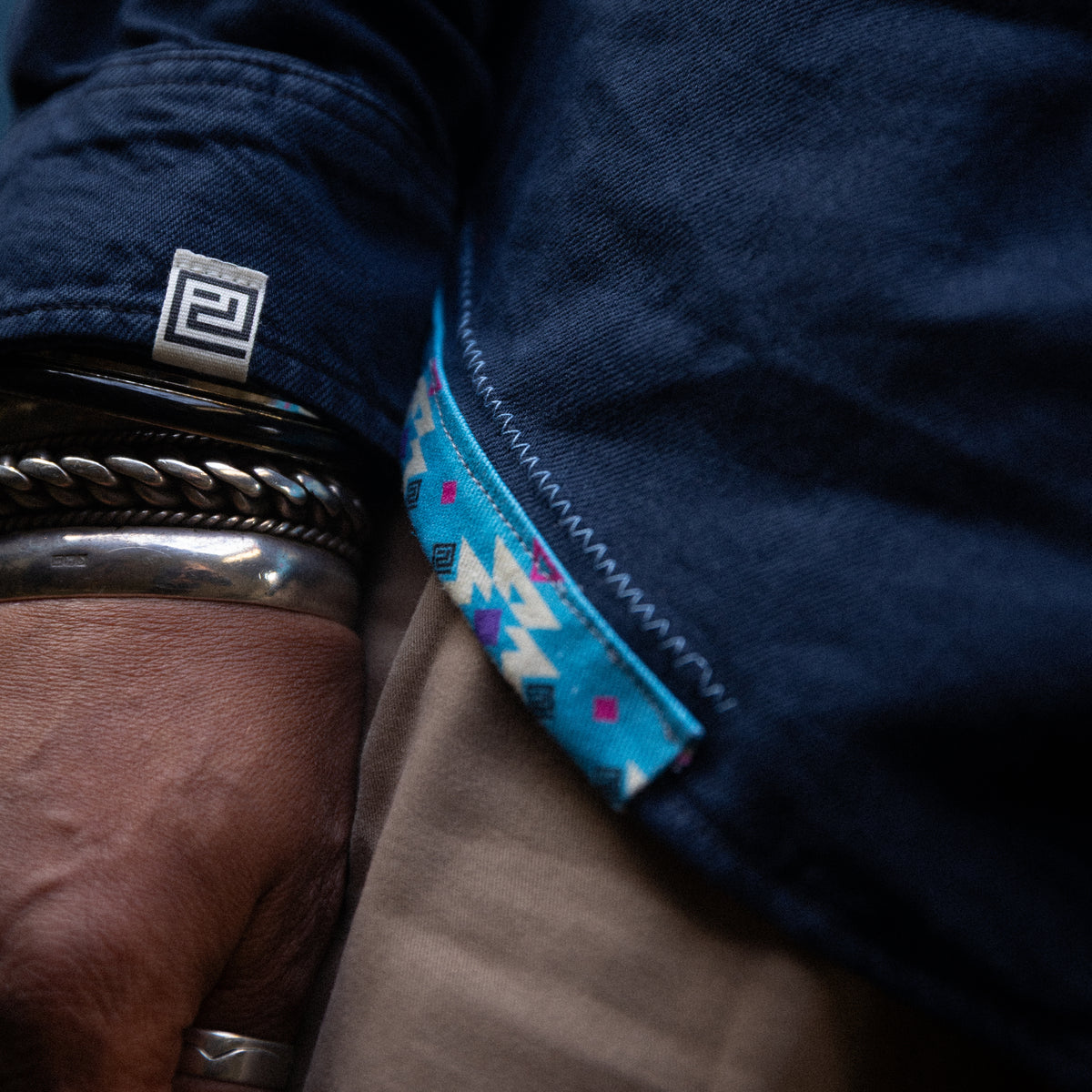 Close-up of a person wearing a dark blue denim jacket, showing the inner cuff with a colorful geometric pattern lining and a small square logo patch. The person also wears silver bracelets and a ring.