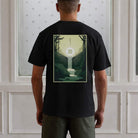 Person wearing a black t-shirt with a nature-themed design on the back, standing against a white paneled wall.