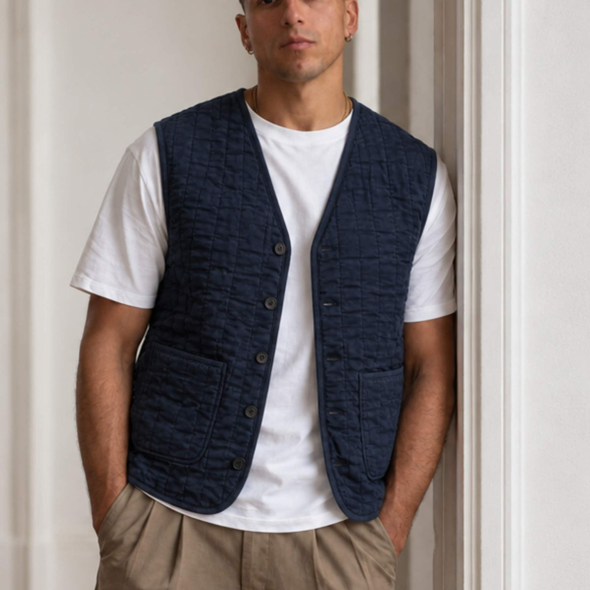A man wearing a navy blue quilted paradaiza button-up vest over a white t-shirt and khaki pants, standing in a doorway.