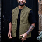 A man wearing a paradaiza  olive green zip-up utility vest over a black t-shirt, standing in front of a dark wooden door.
