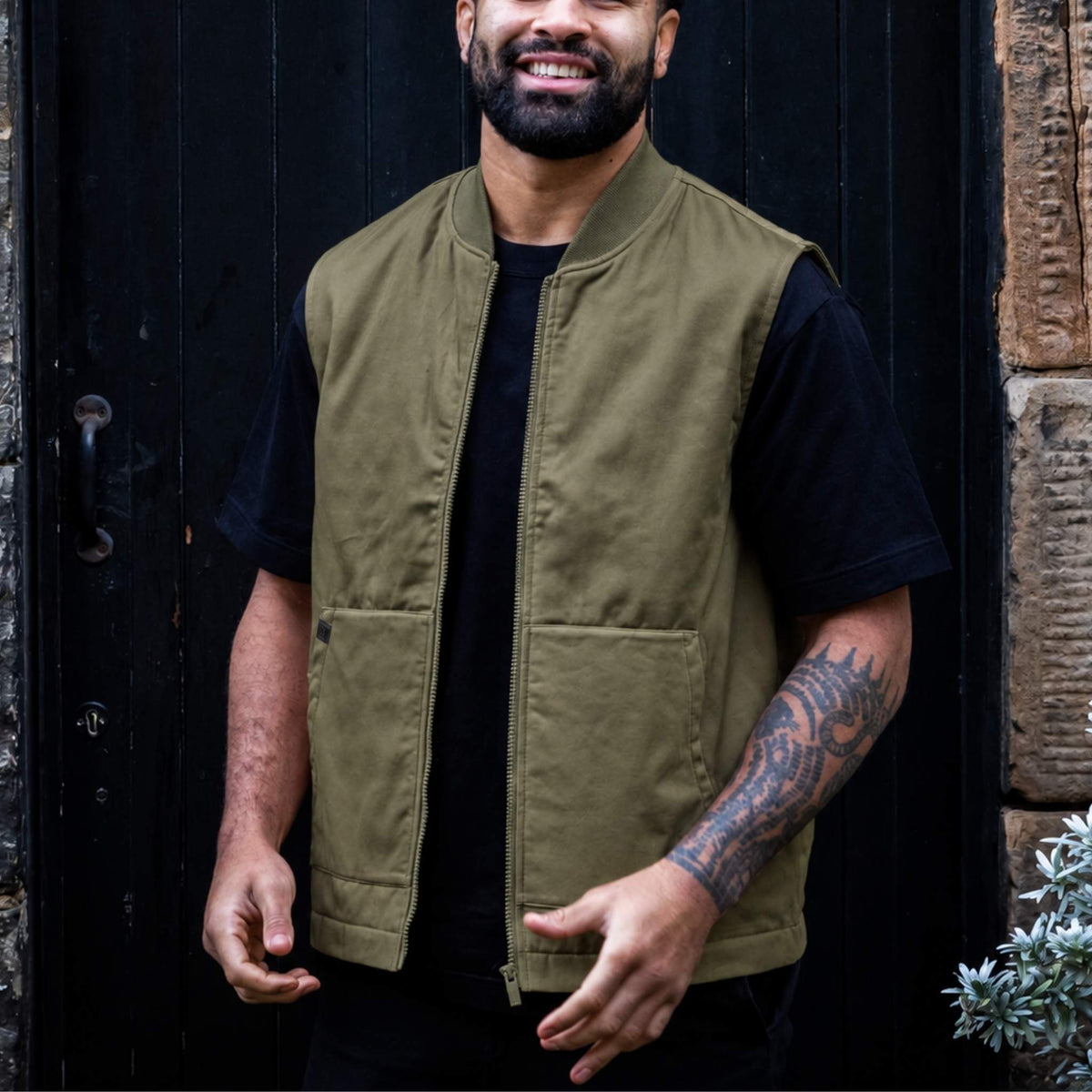 A man wearing a paradaiza  olive green zip-up utility vest over a black t-shirt, standing in front of a dark wooden door.